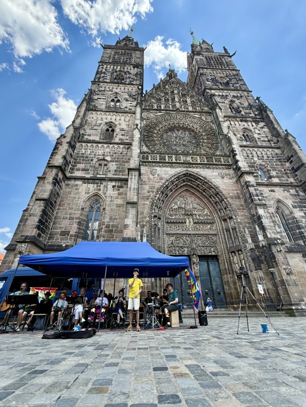 Eine Gruppe von Jugendlichen und Erwachsenen aus der Schülerband Studio D sitzt unter einem blauen Pavillon vor der beeindruckenden Fassade der Lorenzkirche Nürnberg, einer großen gotischen Kirche mit zwei Türmen. Die Personen spielen verschiedene Instrumente wie Trommel, Schlagzeug und Klangstäbe. Im Vordergrund sind Notenständer und Mikrofone zu sehen, im Hintergrund ragt die detailreiche Steinfassade der Kirche vor blauem Himmel mit Wolken auf.