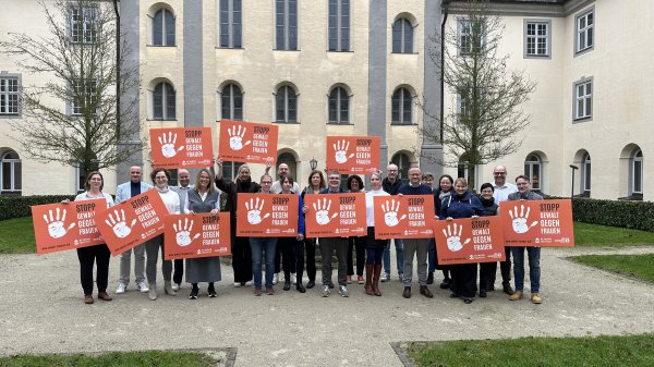 Eine Gruppe Menschen steht vor einem Gebäude und hält orangefarbene Schilder mit der Aufschrift „Stopp Gewalt gegen Frauen“.