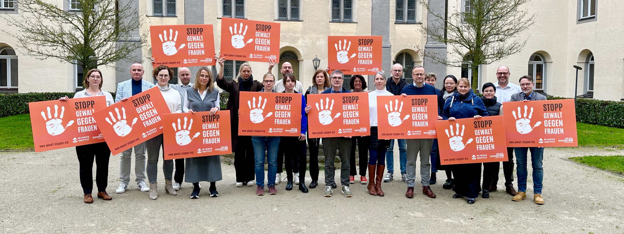 Eine Gruppe Menschen steht vor einem Gebäude und hält orangefarbene Schilder mit der Aufschrift „Stopp Gewalt gegen Frauen“.