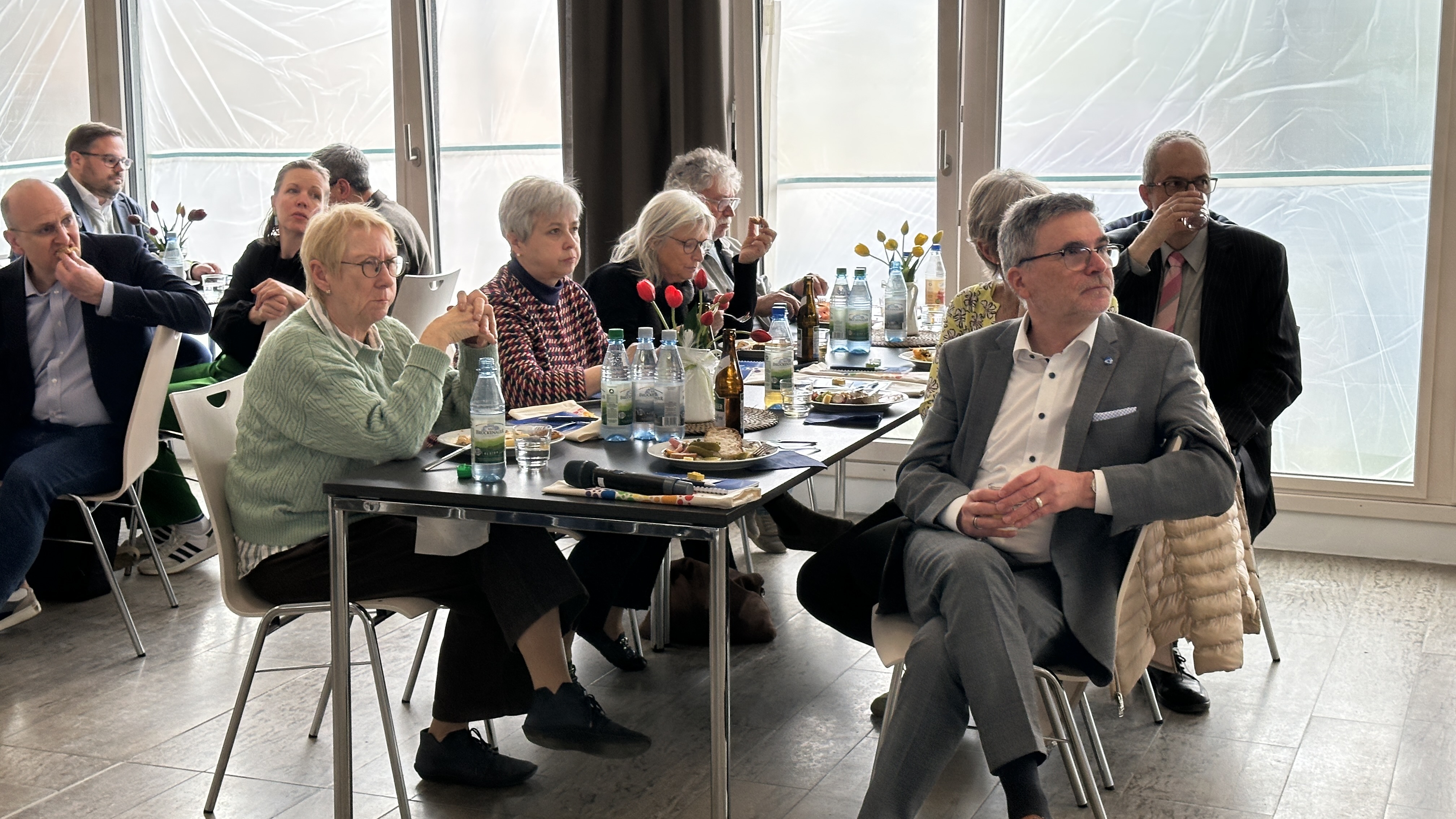 Mehrere Menschen sitzen an Tischen in einem Raum. Sie hören einem Redner aufmerksam zu. 