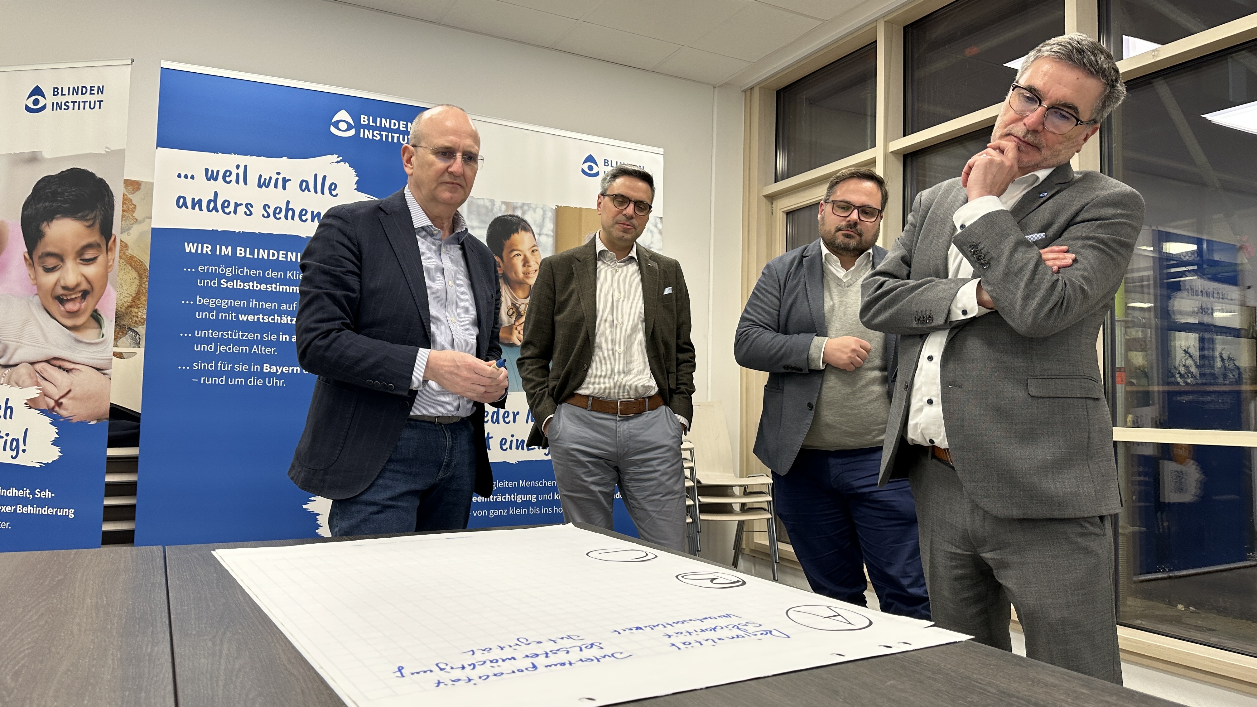 Vier Männer stehen um einen Tisch herum und betrachten ein großes weißes Blatt Papier. Sie überlegen, was sie darauf schreiben sollen. 