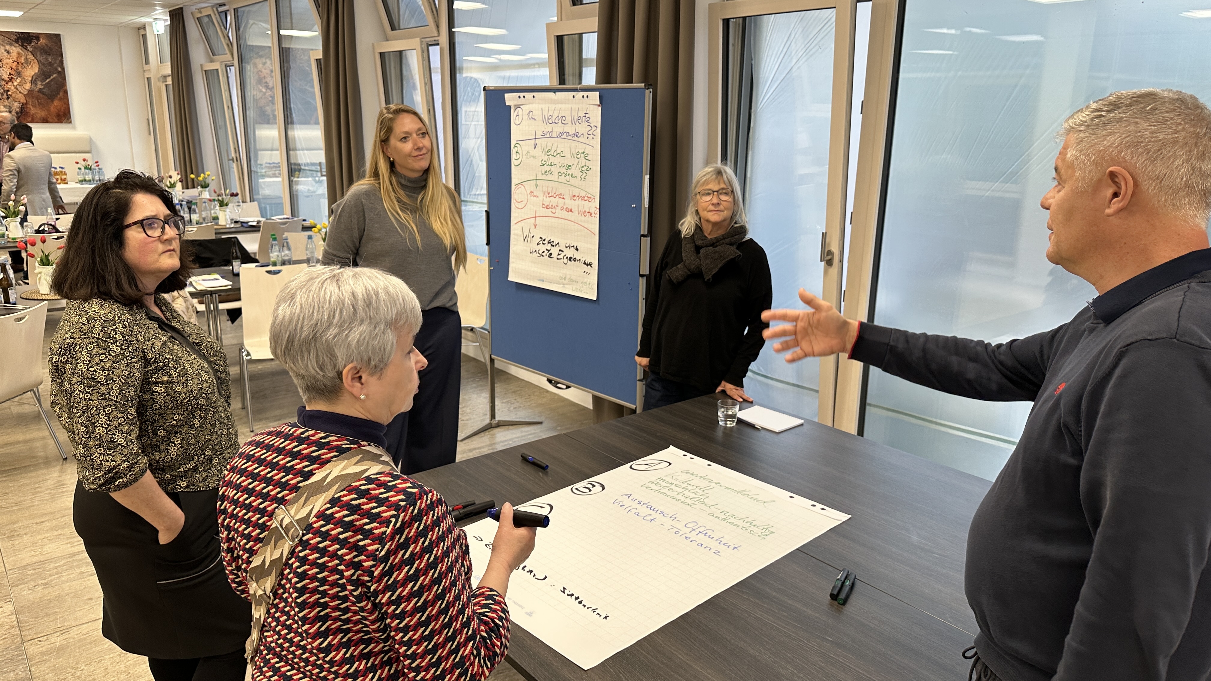 Mehrere Personen stehen um einen Tisch herum und betrachten ein großes weißes Blatt Papier. Sie überlegen und diskutieren, was sie darauf schreiben sollen. 