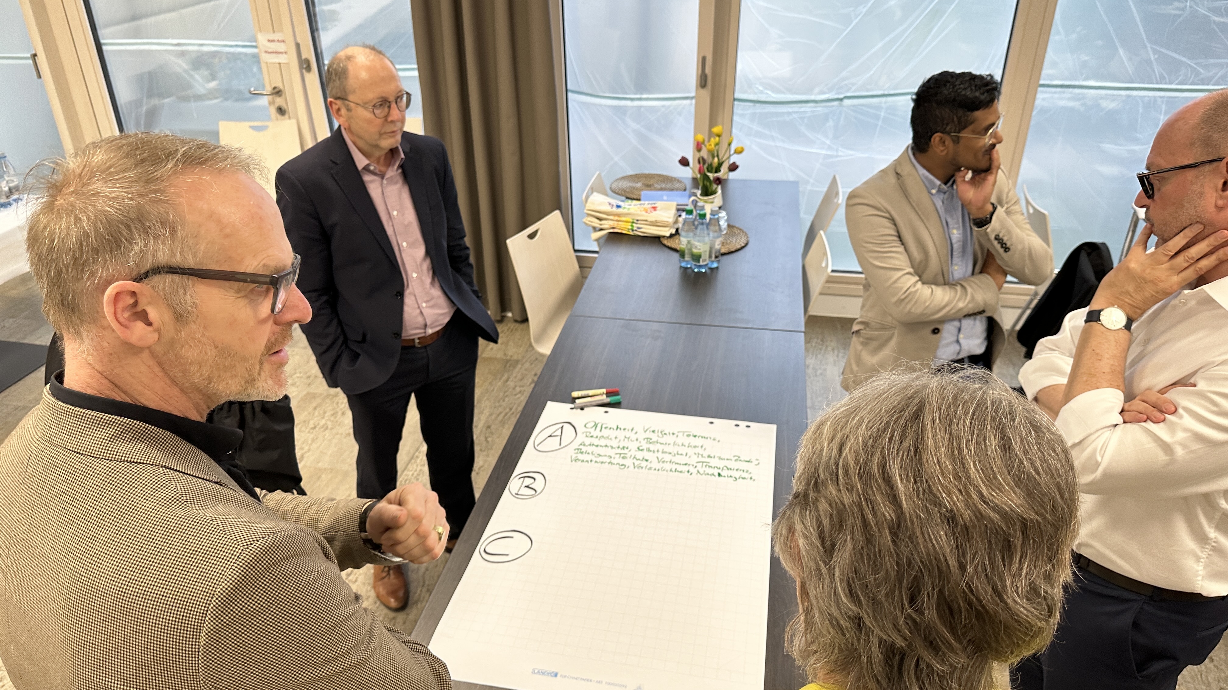 Mehrere Personen stehen um einen Tisch herum und betrachten ein großes weißes Blatt Papier. Sie überlegen und diskutieren, was sie darauf schreiben sollen. 