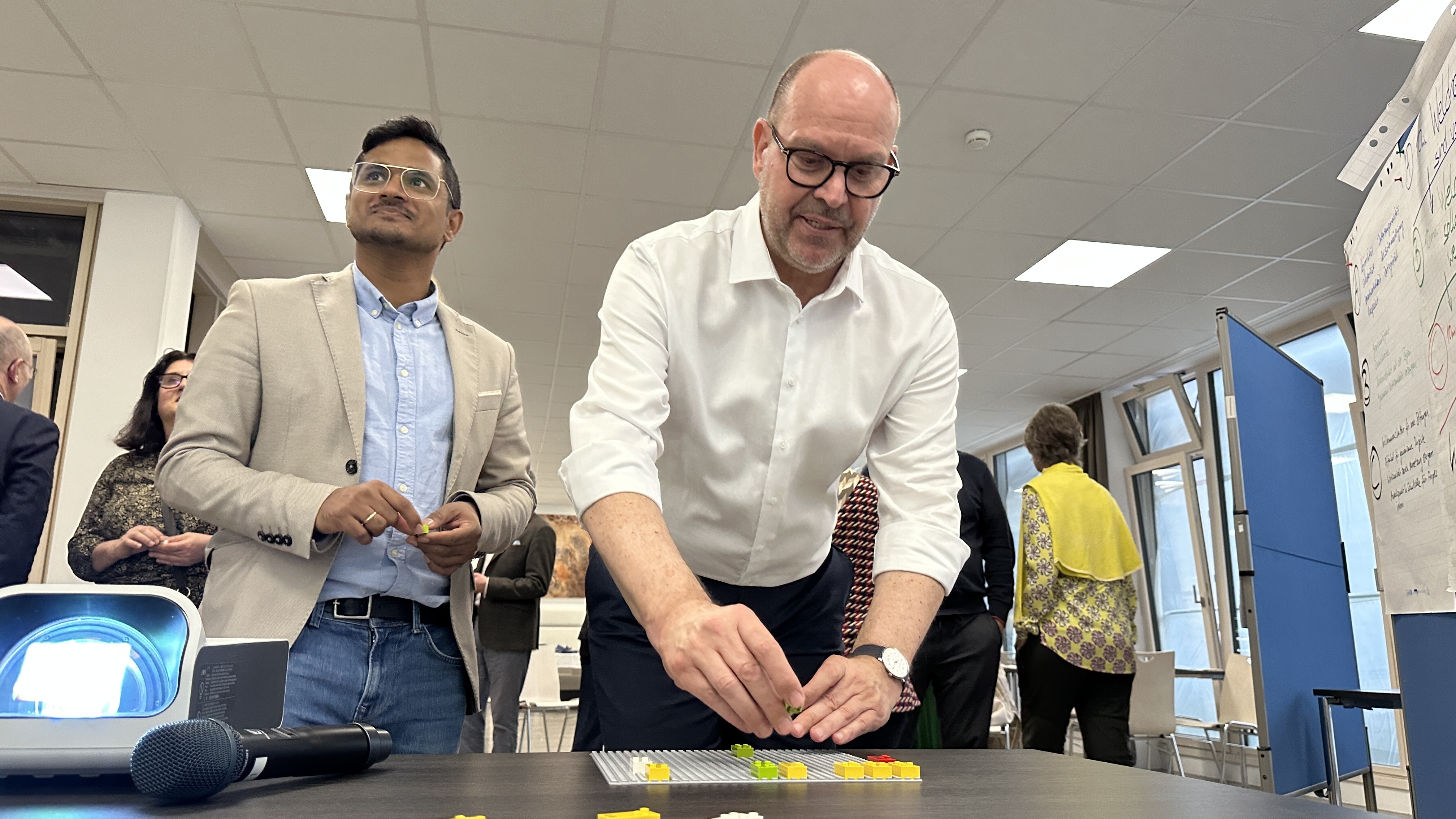 Zwei Männer stehen an einem Tisch. Der Mann vorne legt kleine bunte Bausteine auf eine Platte. Der andere Mann schaut zu und hält ebenfalls ein Teil in der Hand.