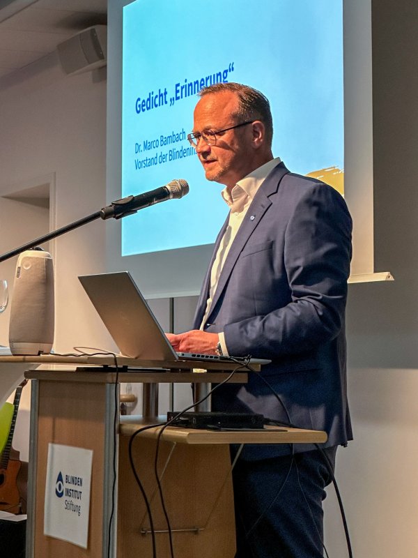 Seitliches Foto von einem Mann im Anzug (Marco Bambach), der vor einem Mikrofon steht und ein Gedicht vorträgt.