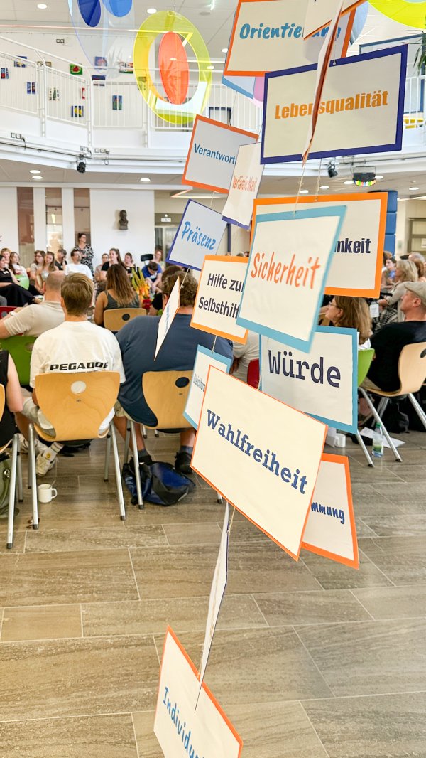 Im Vordergrund: hängende Papiere mit Schriften wie: Wählfreiheit, Sicherheit & Würde und im Hintergrund das Publikum sitzend.