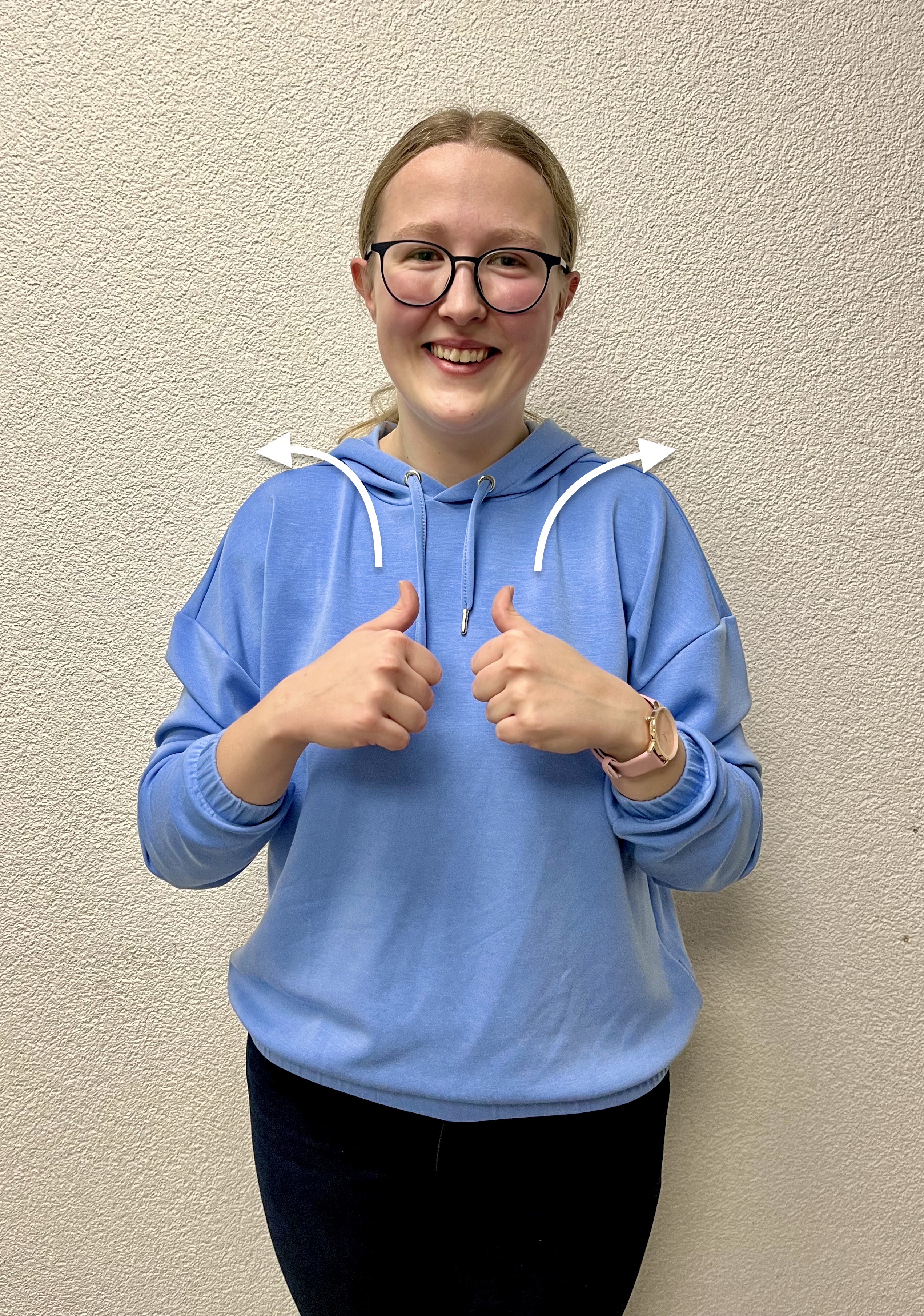 Eine lächelnde junge Frau in einem hellblauen Hoodie hält beide Daumen nach oben. Weiße Pfeile um ihre Schultern heben ihre Gesten hervor.