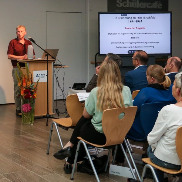 Ein Mann (Prof. Oded Heilbronner) steht in einem dunkelroten Hemd vor dem Publikum und hält eine Präsentation über die Zwangssterilisation während der NS Zeit