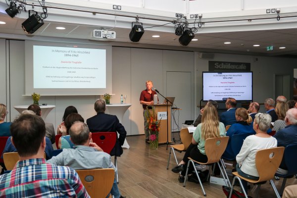 Ein Mann (Prof. Oded Heilbronner) steht in einem dunkelroten Hemd vor dem Publikum und hält eine Präsentation über die Zwangssterilisation während der NS Zeit . Das Publikum ist im zugerichtet und hört ihm zu.