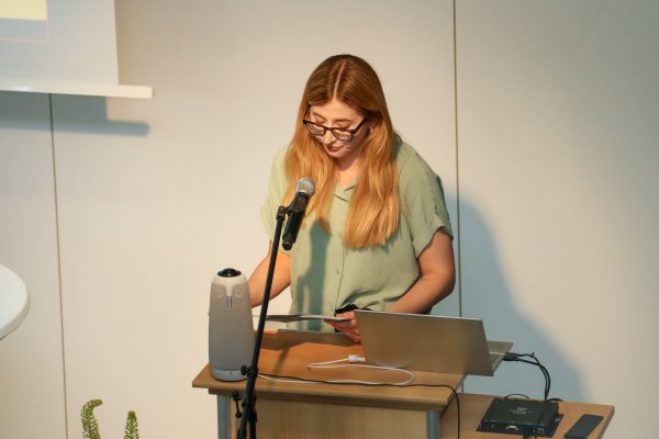 Eine Frau (Amelie Seidl vom Blindeninstut) steht vor einem Mirkofon und übersetzt den Votrag von Prof. Oded Heilbronner ins Deutsche.