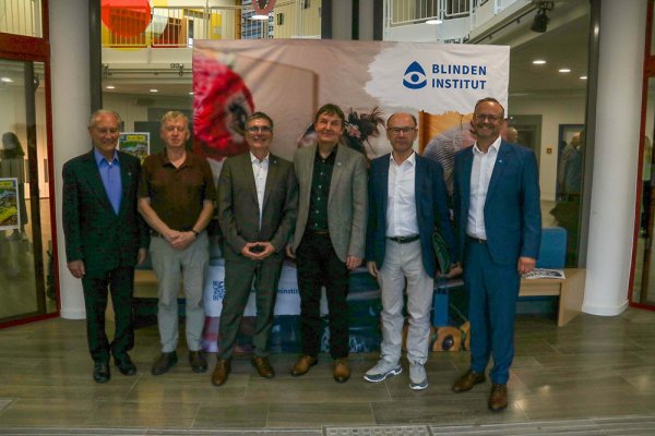 Gruppenfoto von sechs Männern in Anzügen, die vor einem großen Blindeninstitut Banner stehen
