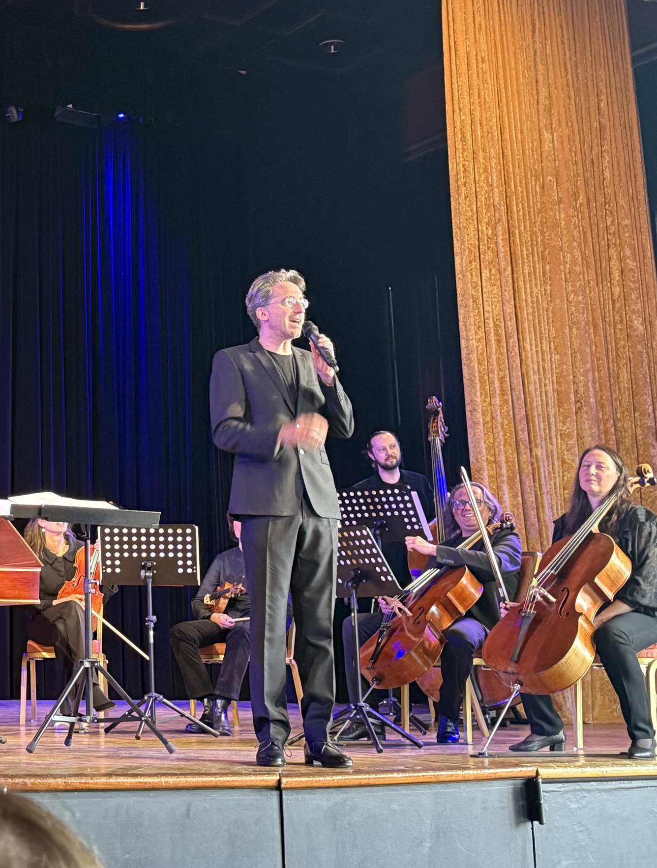 auf dem Bild sieht man einen Mann mit einem Mikrofon in der Hand. Im hintergrund sitzen Cellisten.