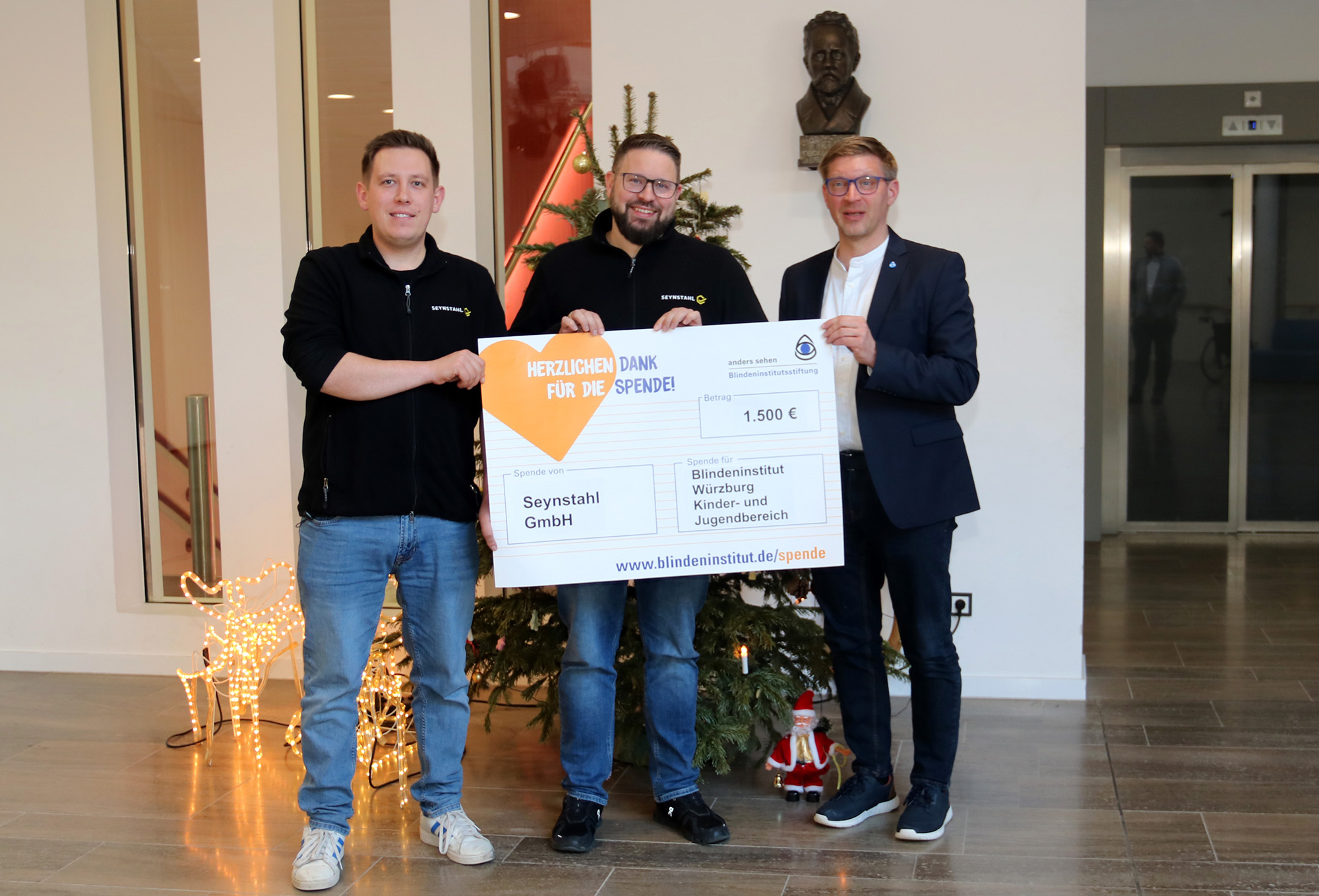 Drei Männer stehen vor einem Weihnachtsbaum und halten einen Spendenscheck in der Hand