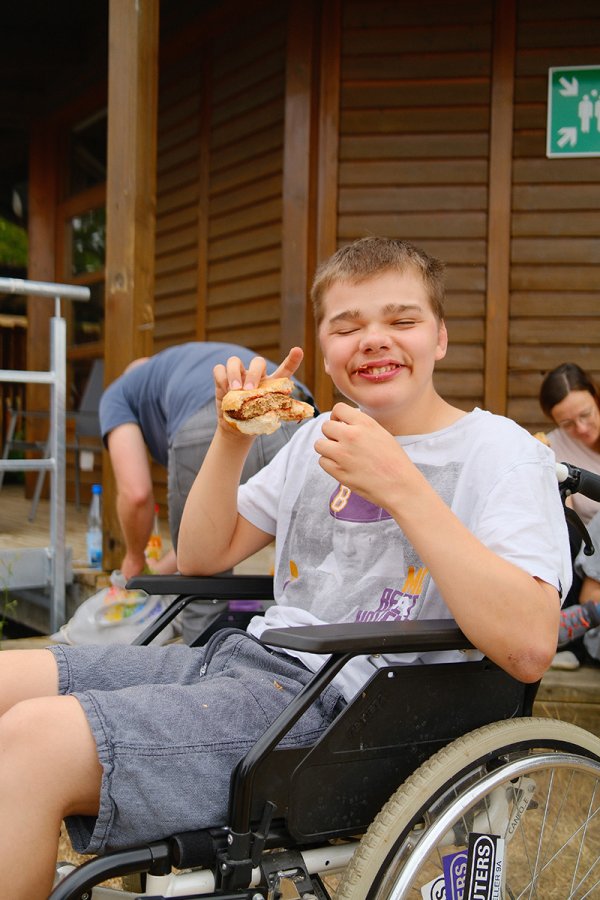 Ein Junge im Rollstuhl mit einem Grillsteak im Brötchen lacht in die Kamera.