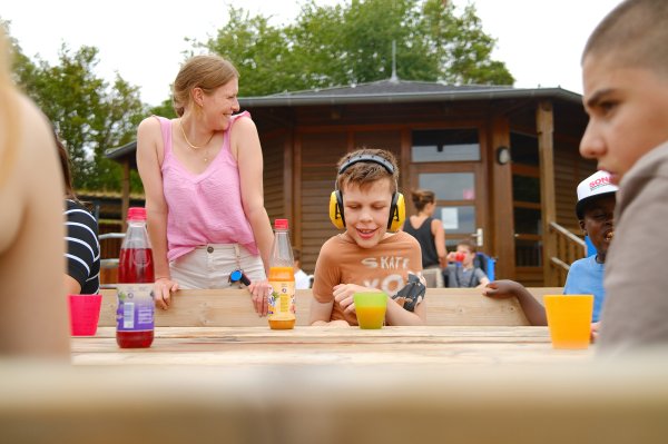 Drei Kinder und eine junge Frau sitzen an einem Holztisch. Ein junge schenkt sich Saft ein.