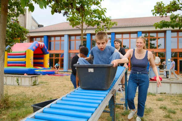 Ein Junge fährt in einer Plastikwanne auf einer schrägen Rampe hinunter. Eine junge Frau hält ihn und läuft neben her.