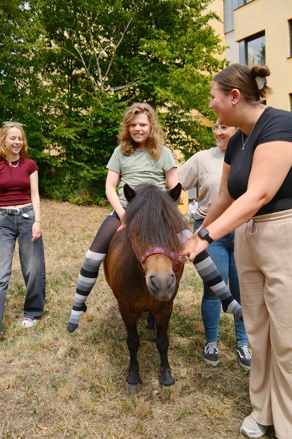 Ein junges Mädchen reitet auf einem Pony und lacht in die Kamera. Eine Frau steht rechts neben ihr und hält sie.