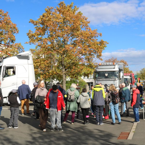 Eine große Gruppe Menschen steht im Halbkreis an einer Straße und sieht mehreren großen LKW-Zugmaschinen zu, die vorbeirollen.