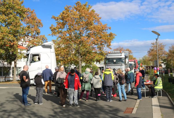 Eine große Gruppe Menschen steht im Halbkreis an einer Straße und sieht mehreren großen LKW-Zugmaschinen zu, die vorbeirollen.