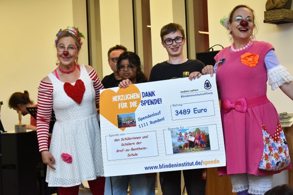 Rechts und links neben einem großen Spendenscheck stehen zwei Clowninnen und halten den Scheck an der Seite. Hinter dem Scheck stehen ein Erwachsenener und zwei Schüler*innen.