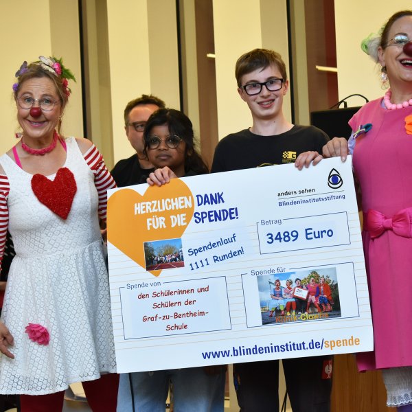 Rechts und links neben einem großen Spendenscheck stehen zwei Clowninnen und halten den Scheck an der Seite. Hinter dem Scheck stehen ein Erwachsenener und zwei Schüler*innen.
