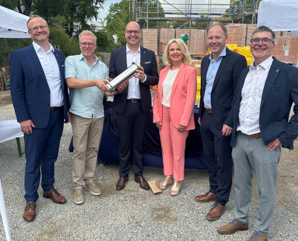 Fünf Männer und eine Frau stehen im Halbkreis auf einer Baustelle. Zwei Männer in der Bildmitte halten einen Edelstahlbehälter in der der Hand.
