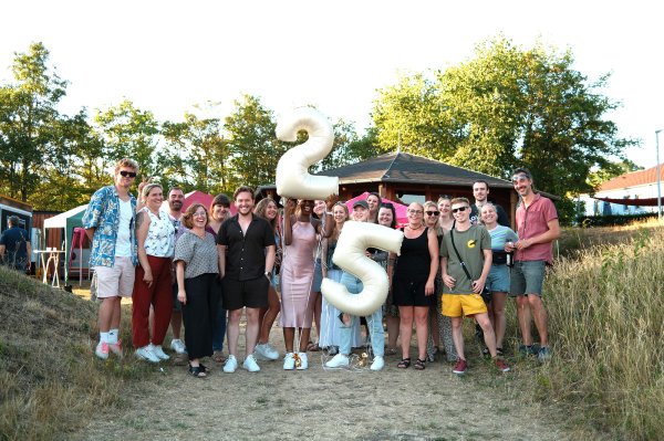 Eine Gruppe vom Menschen steht im Halbkreis in einer Wiese. Zwei Personen in der Mitte halten je einen Ballon in Form einer Zwei und einer Fünf.