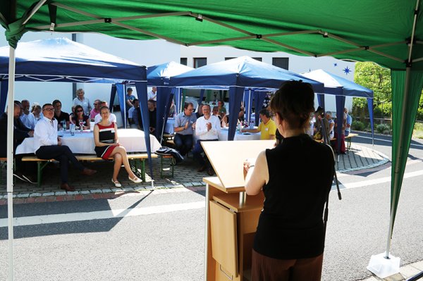 Im Vordergrund steht Dr. Anja Klafke mit dem Rücken zum Betrachter vor einem Rednerpult. Im Bildhintergrund sitzen die Gäste der Jubiläumsveranstaltung auf Bierzeltgarnituren unter Pavillons und hören der Ansprache zu.