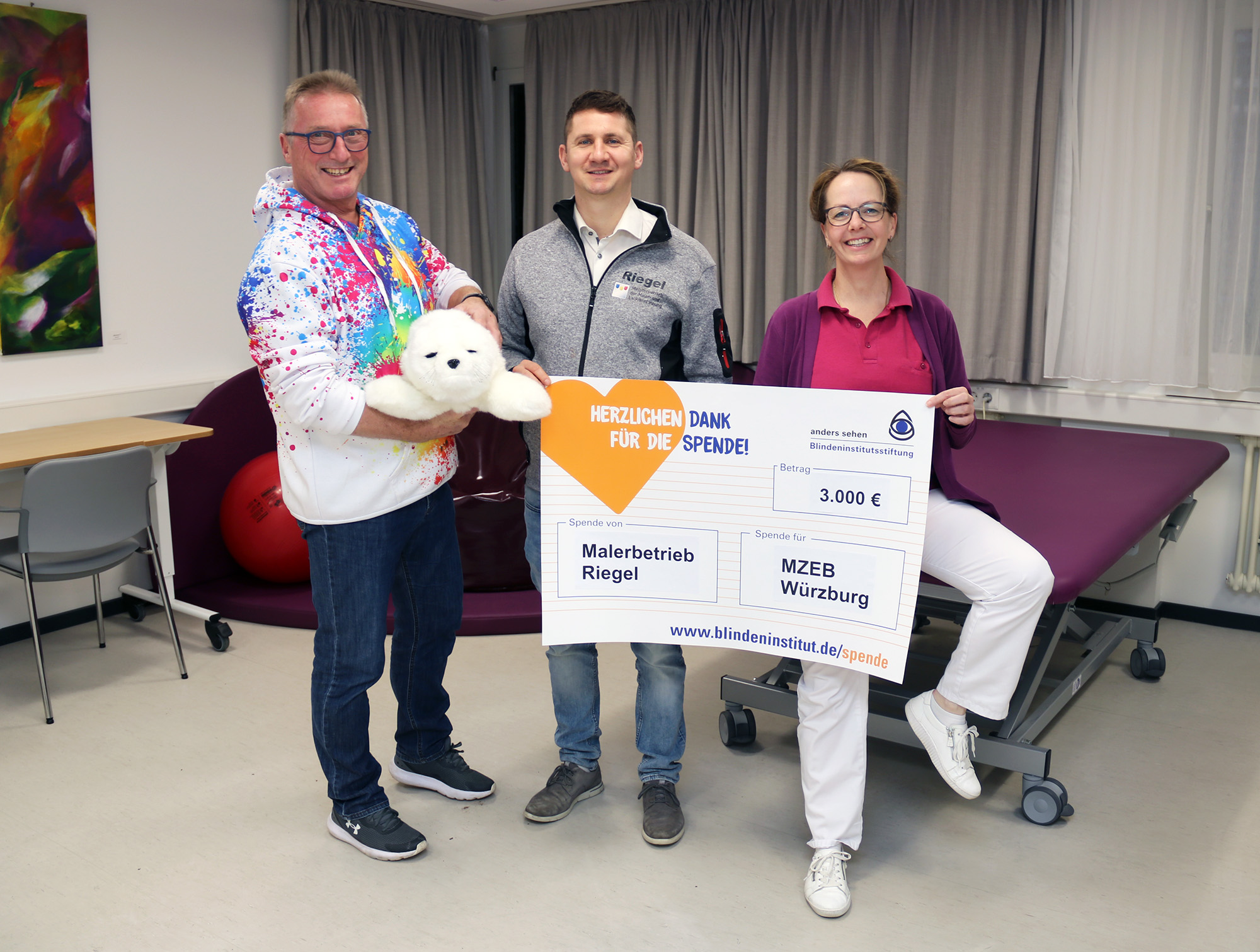 Zwei Männer und eine Frau stehen in einem Behandlungszimmer. Der linke Mann hält eine kleine Stoffrobbe in der Hand. Der mittlere Mann und die Frau halten einen großen Spendenscheck in den Händen.