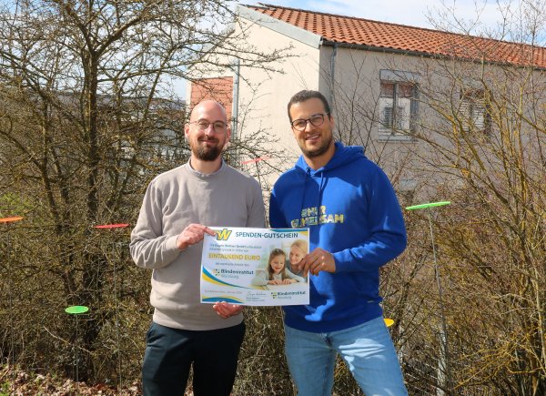 Zwei Männer stehen auf dem Außengelände vor einem Hau s und halten einen Spendenscheck in der Hand.