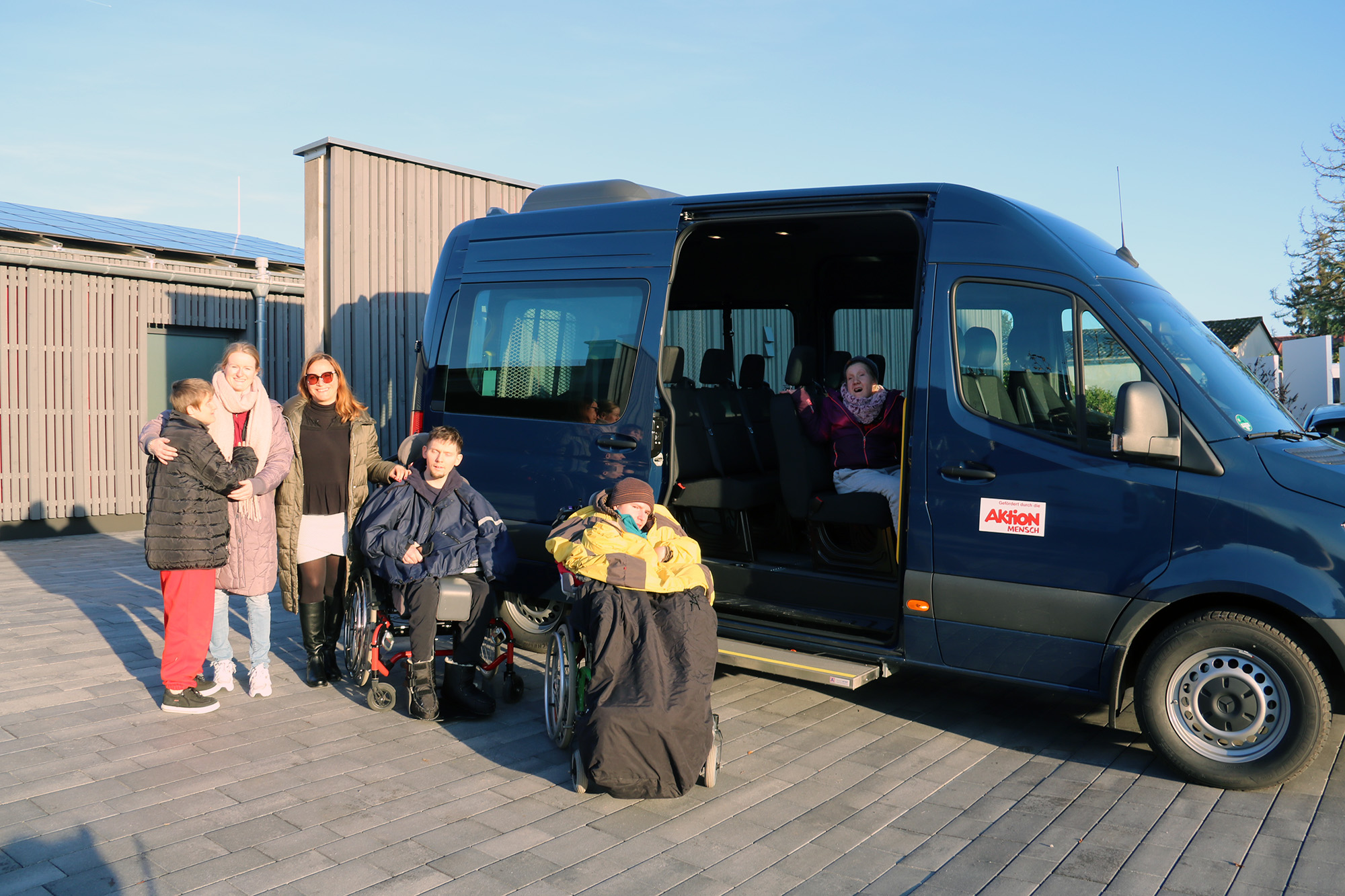 Fünf Erwachsene, davon zwei Rollstuhlfahrer, stehen vor einem neuen Kleinbus, eine Frau sitzt im Bus, dessen Schiebetür geöffnet ist.