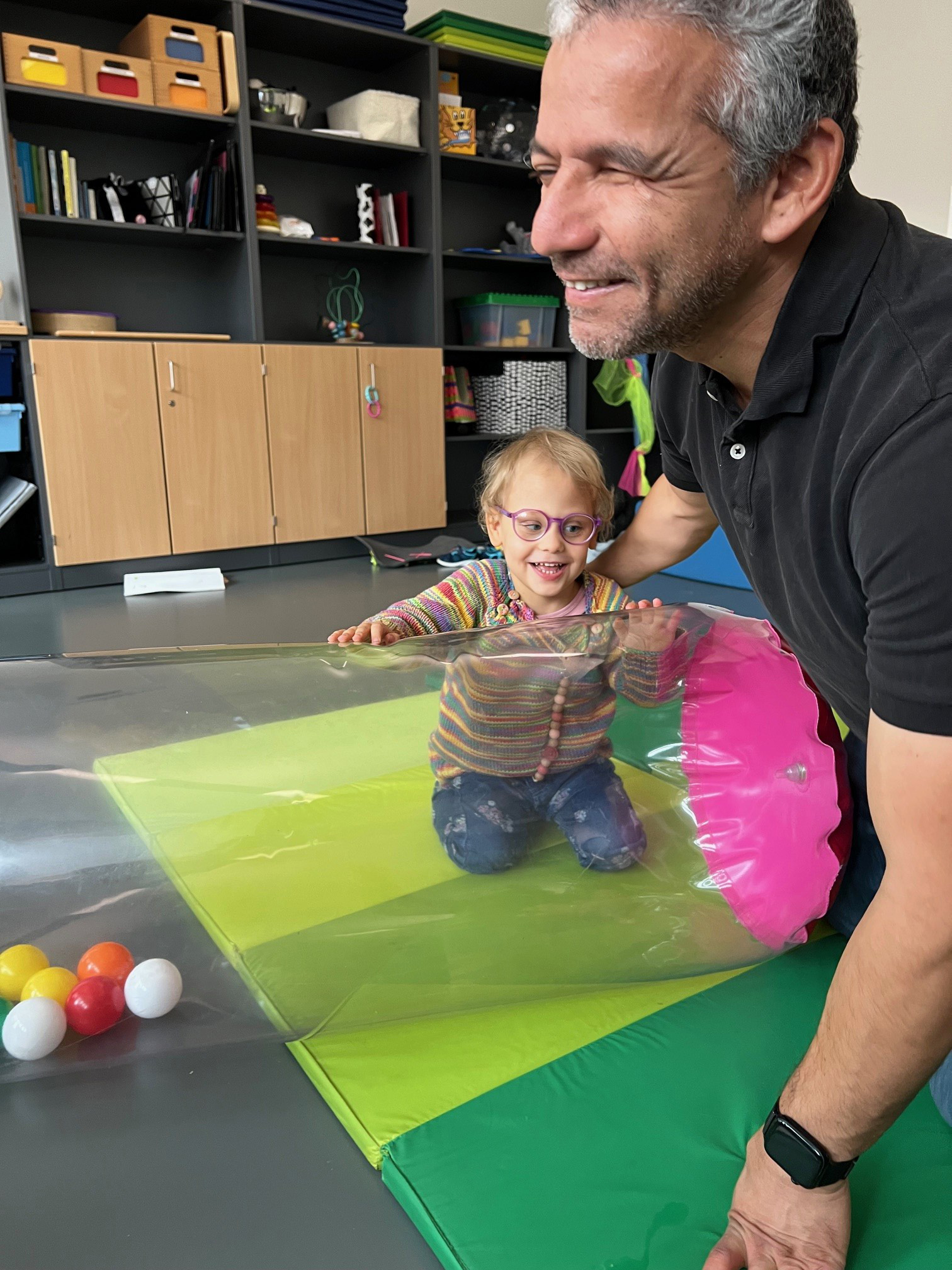 Ein Mann kniet neben einem Kleinkind, das auf einer aufblasbaren Rolle liegt.