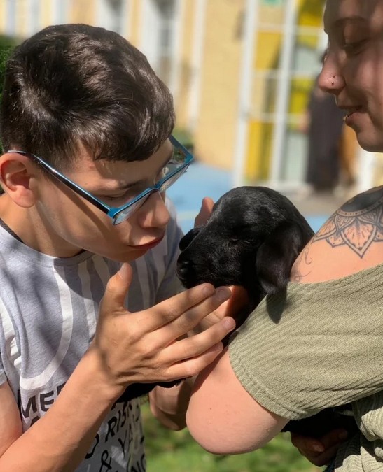 Das Bild zeigt einen Jungen, der ein schwarzes Lämmchen in den Händchen hält