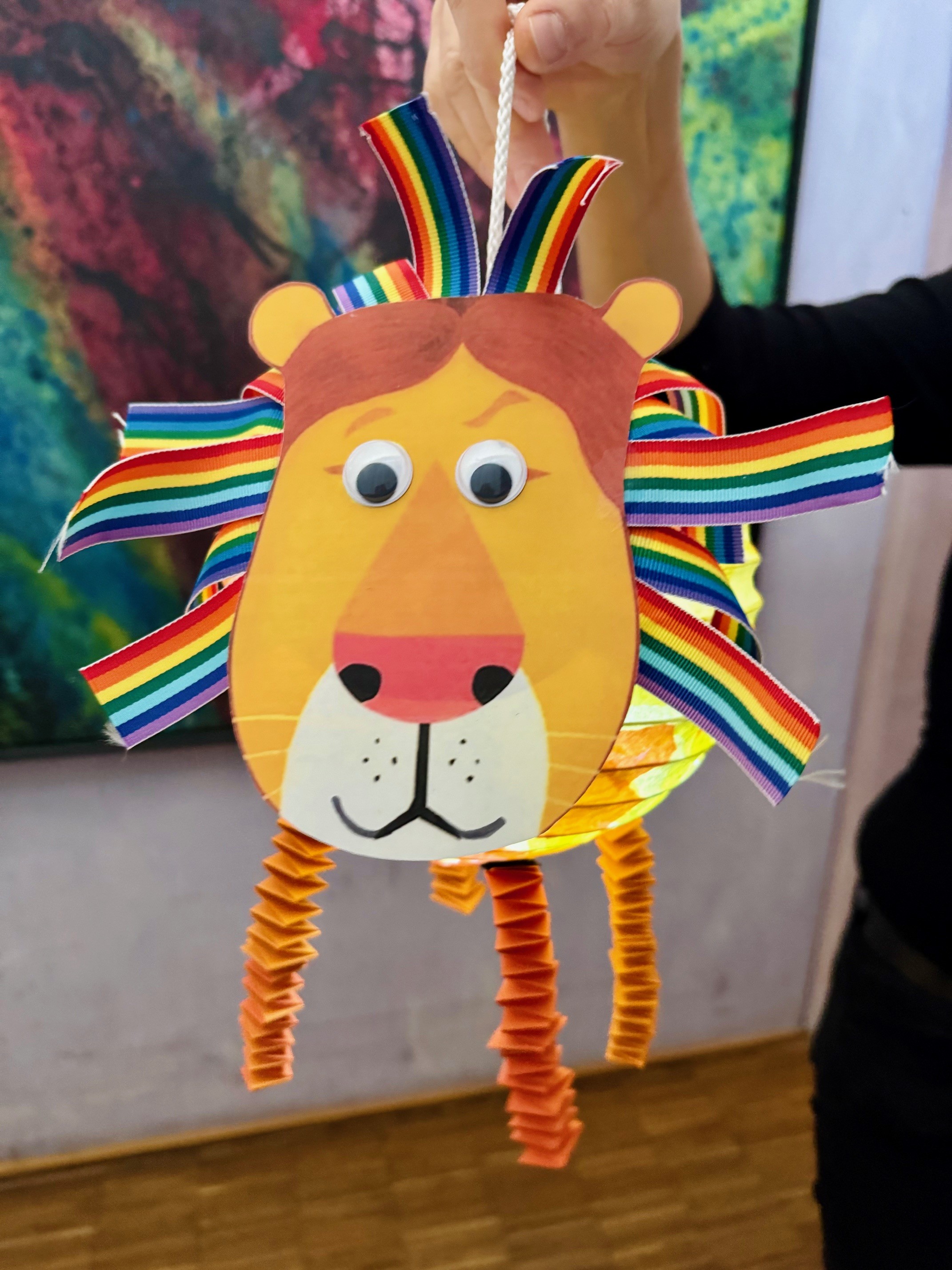 Es ist eine sehr bunte Löwenlaterne zu sehen. Sie hat kräftige gelbe und orangefarbene Akzente. Eine wilde Regenbogenmähne und wartet auf ihren Einsatz um hell in der dunklen Aula zu strahlen.
