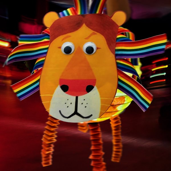 Das Bild zeigt eine Laterne aus Tonpapier in Gestalt eines Löwenkopfs. Die Löwenmähne besteht aus Bändern in Regenbogenfarben. Der Löwe hat eine große Nase, eine weiße Zeichnung unter seinem Maul und lustige Wackelaugen, die ihn sehr freundlich aussehen lassen.