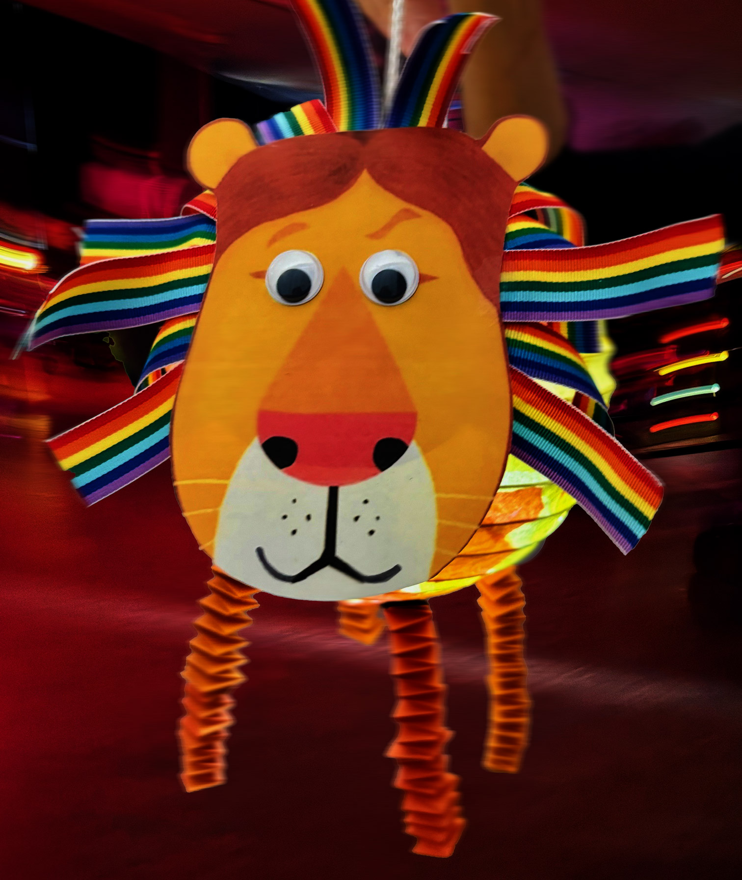 Das Bild zeigt eine Laterne aus Tonpapier in Gestalt eines Löwenkopfs. Die Löwenmähne besteht aus Bändern in Regenbogenfarben. Der Löwe hat eine große Nase, eine weiße Zeichnung unter seinem Maul und lustige Wackelaugen, die ihn sehr freundlich aussehen lassen.
