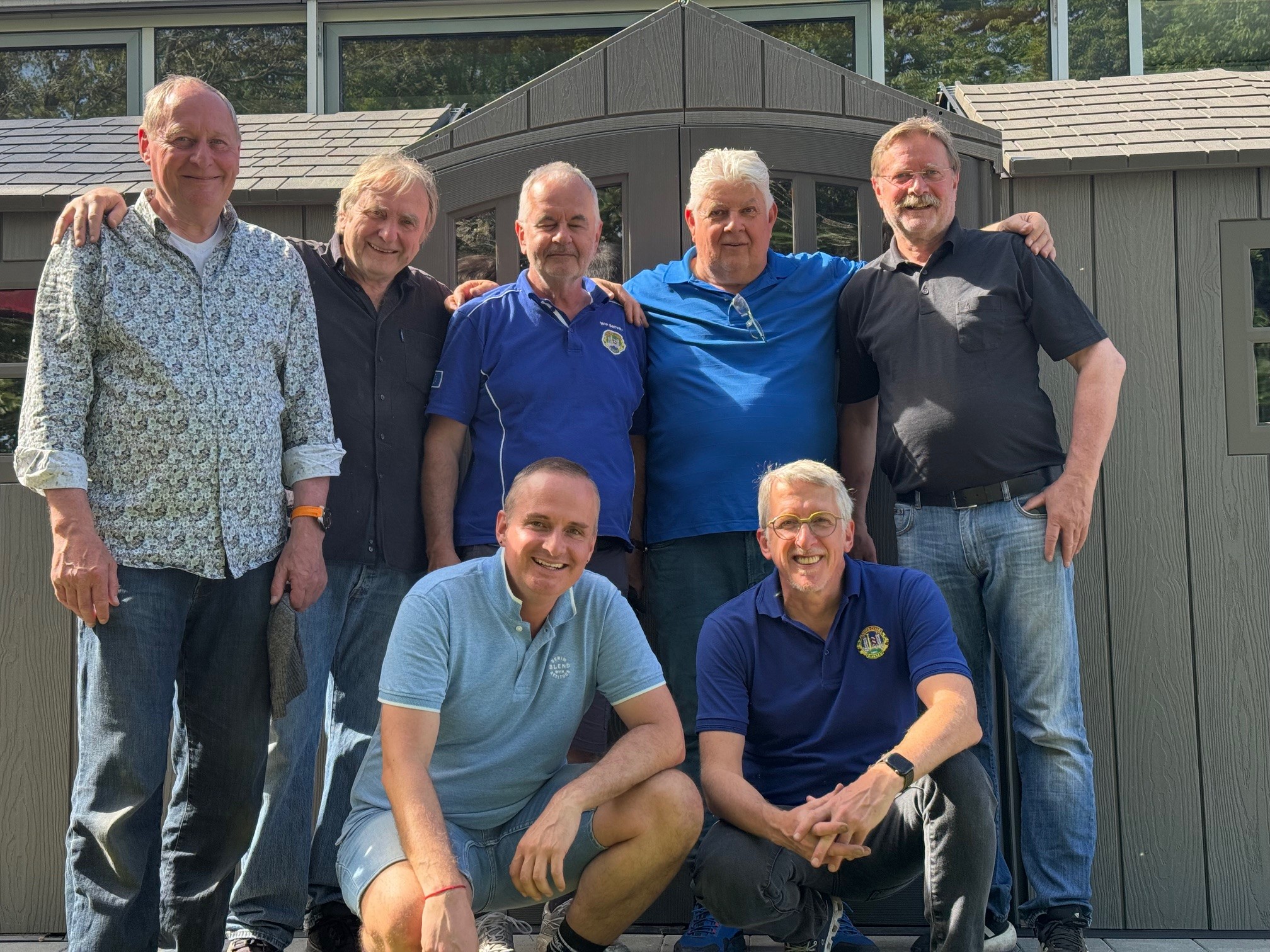 Sieben Mitglieder des Lions Club stehen und knien gemeinsam mit dem Insitutitsleiter Dominic Weufen vor dem neu errichteten grauen Fahrradschuppen auf einem Außengelände. Die Lions haben den Fahrradschuppen gemeinsam aufgebaut. Alle Personen tragen Freizeitkleidung, aufgenommen bei Tageslicht.