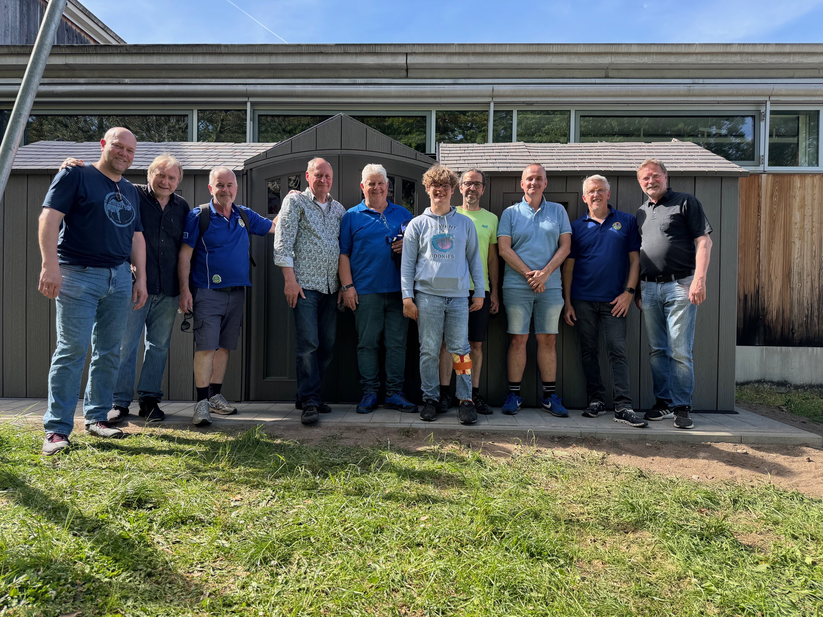 Die beteiligten Mitglieder des Lions Club stehen und knien gemeinsam mit dem Insitutitsleiter Dominic Weufen und weiteren Helfern des Blindeninstituts  vor dem neu errichteten grauen Fahrradschuppen auf einem Außengelände. Die Lions haben den Fahrradschuppen gemeinsam aufgebaut. Alle Personen tragen Freizeitkleidung, aufgenommen bei Tageslicht.