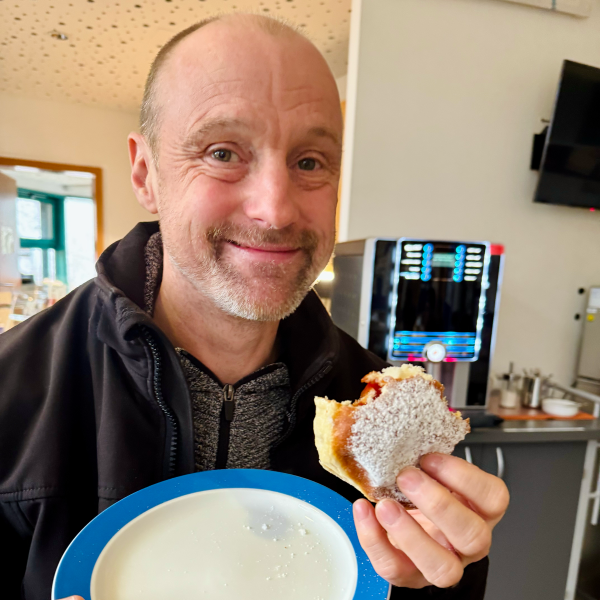 Ein gutgelaunter, lächelnder Mitarbeiter des Blindeninstituts Rückersdorf hält einen leckeren, angebissenen Krapfen in der Hand.