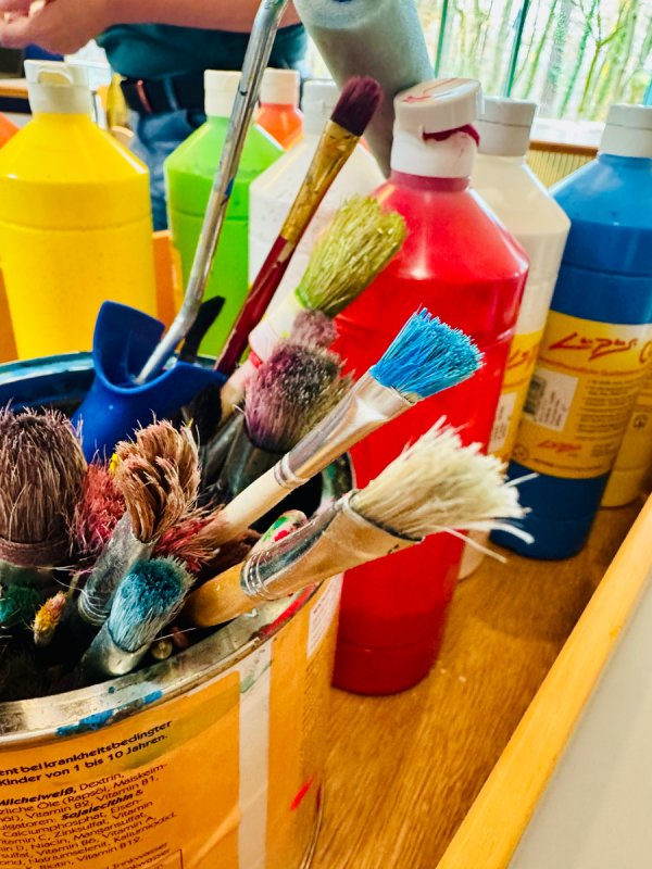 Auf dem Bild sieht man einen Becher voller benutzter Pinsel, welche durch Farbreste bunt erscheinen, sowie eine Auswahl an Acrylfarben in der Flasche.
