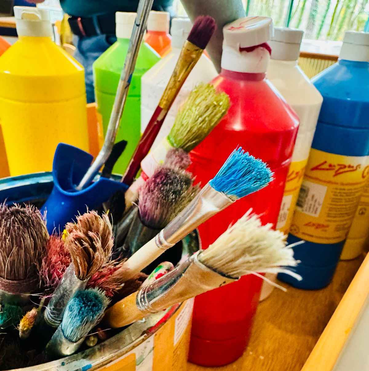 Auf dem Bild sieht man einen Becher voller benutzter Pinsel, welche durch Farbreste bunt erscheinen, sowie eine Auswahl an Acrylfarben in der Flasche.