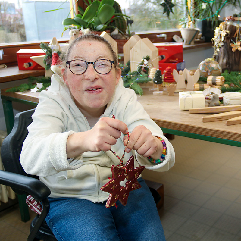 Eine Frau mit Brille sitzt an einem Arbeitstisch und hält einen roten, sternförmigen Weihnachtsschmuck in den Händen; auf dem Tisch liegen Holzarbeiten und weihnachtliche Dekorationen.