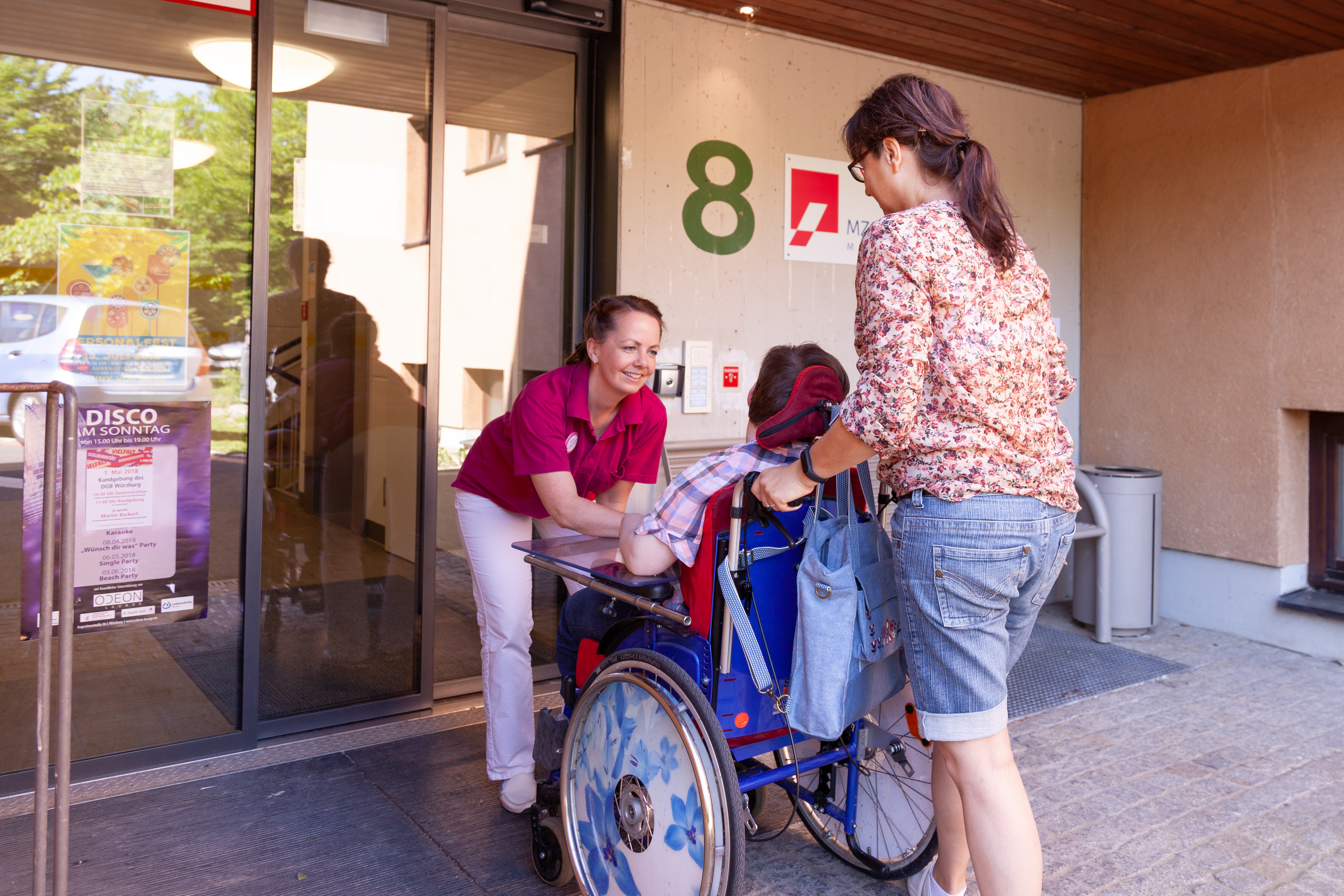 Eine lächelnde Ärztin hilft einer Frau, die im Rollstuhl sitzt, eine andere im Rollstuhl sitzende Person durch die Tür zu schieben.