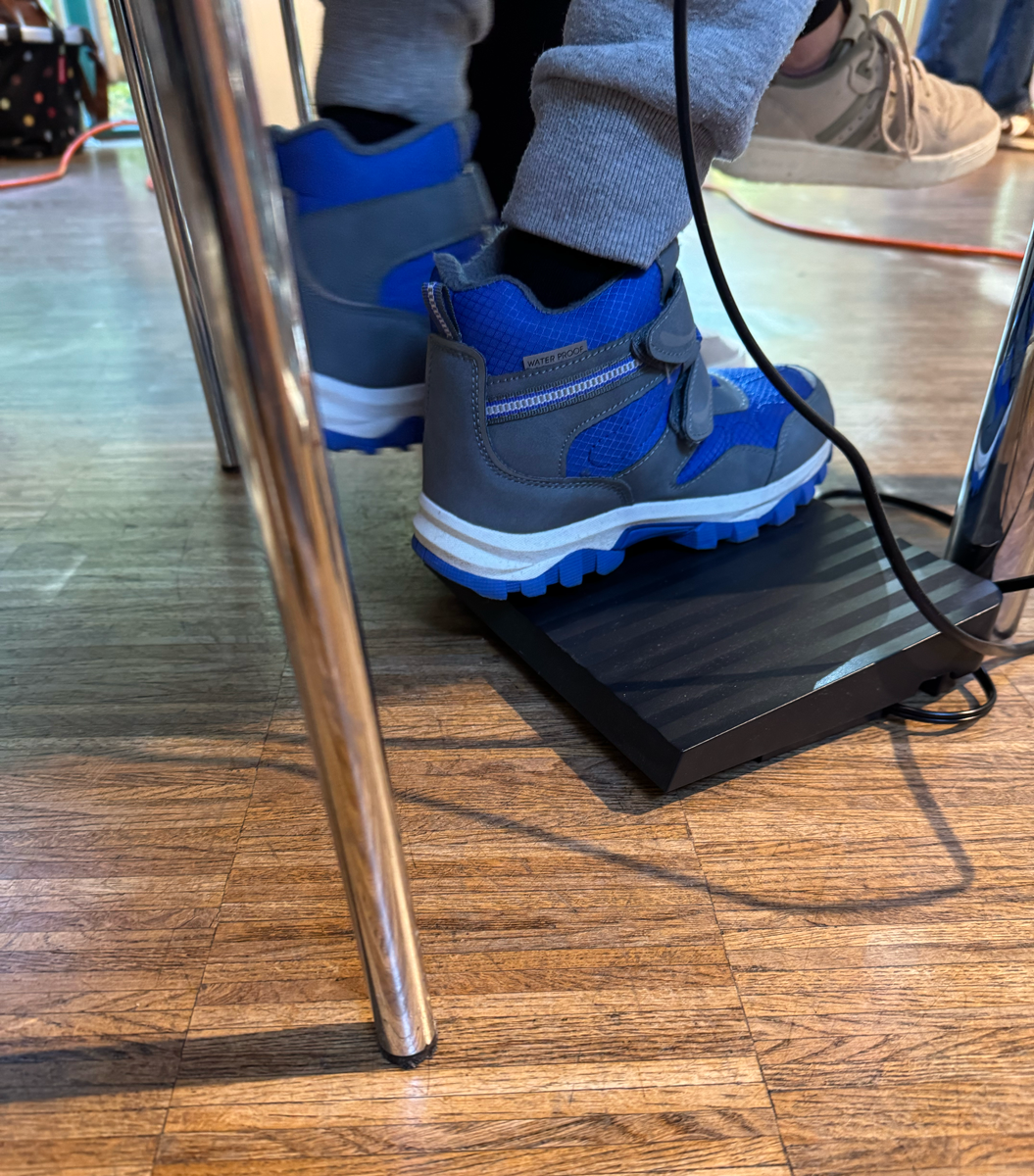 Es ist das Fußpedal einer Nähmaschine zu sehe, das von einem Kind mit blauen Winterstiefeln bedient wird.