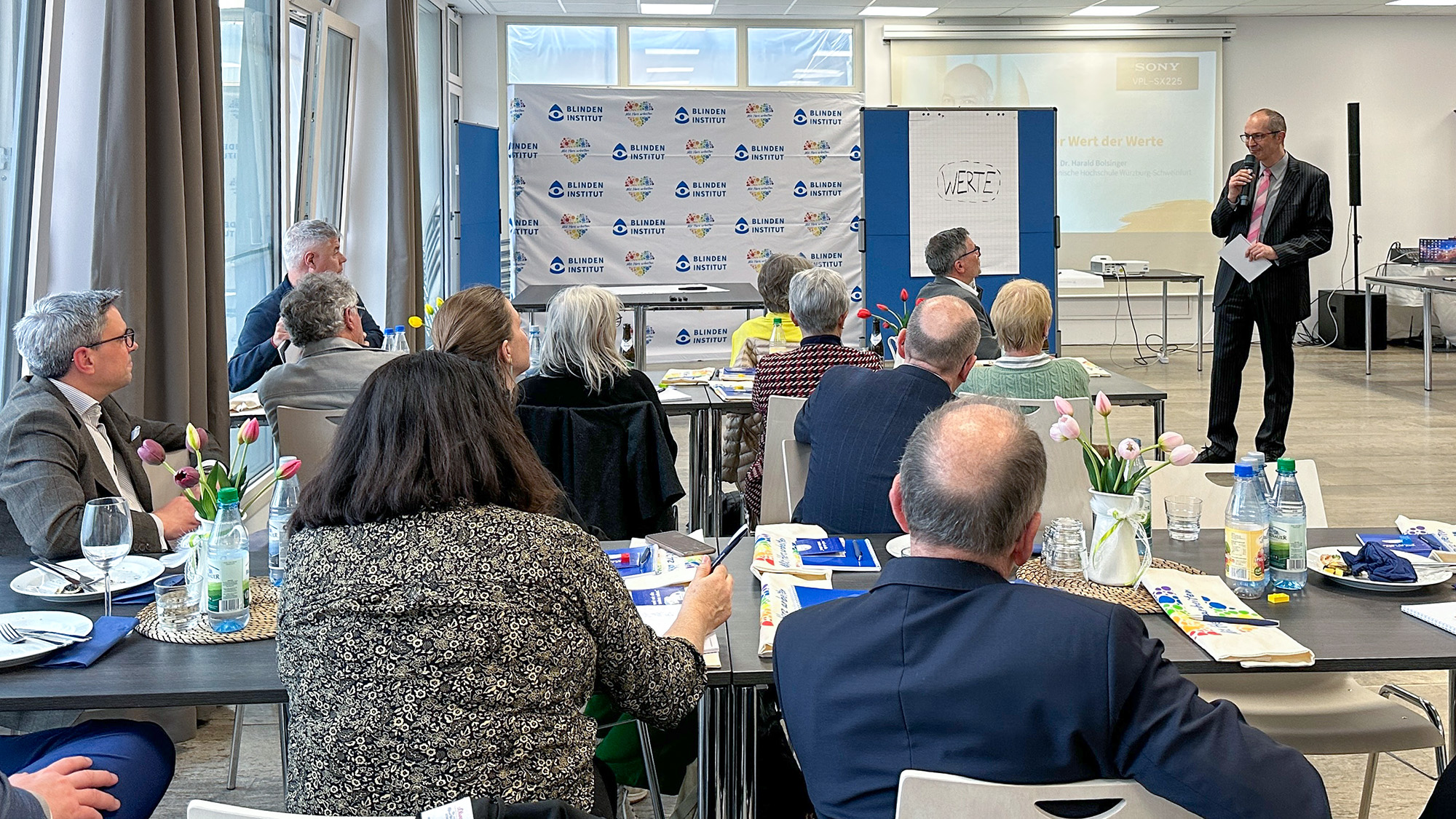 Ein Mann steht vorne im Raum und spricht zu einer Gruppe von Menschen. Die Menschen sitzen an Tischen und hören zu. Auf einer Tafel steht das Wort Werte. Im Hintergrund ist eine Präsentation zu sehen.