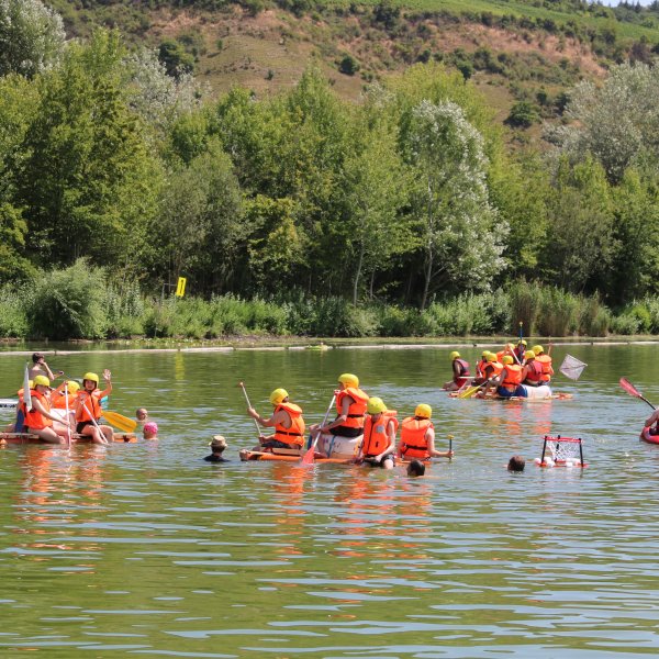 Vier Flöße mit je vier bis fünf Schüler*innen in Schwimmwesten und mit Helmen paddeln in der Mitte eines Badesees und haben viel Spaß.