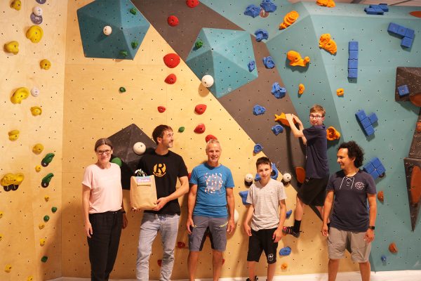 Eine Gruppe mit vier Erwachsenen und zwe Kindern steht vor einer Boulderwand. Ein Kind befindet sich rund einen Meter über dem Boden in der Kletterwand.
