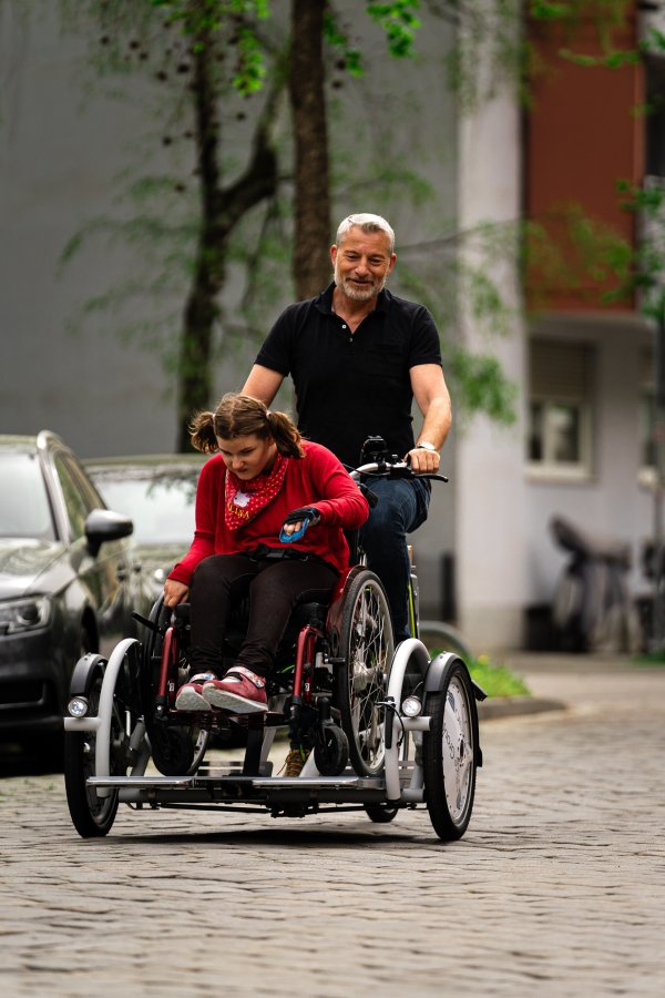 Das Bild zeigt ein Mädchen in einem Rollstuhl. Dieser steht auf einem Rollstuhlfahrrad, das von einem Mann gefahren wird.