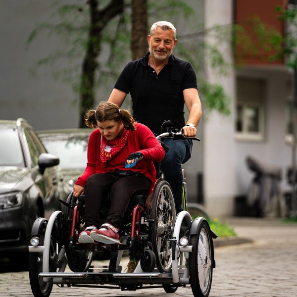 Das Bild zeigt ein Mädchen in einem Rollstuhl. Dieser steht auf einem Rollstuhlfahrrad, das von einem Mann gefahren wird.