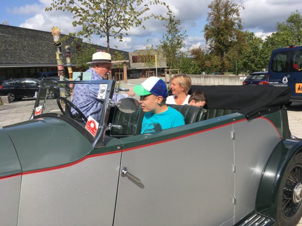 In einem Oldtimer-Cabrio sitzt bei Sonnenschein ein Junge am Steuer. Auf der Rückbank befindet sich eine Frau, während ein Mann in das Cabrio hineinschaut.